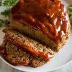 A loaf of meatloaf on a white plate with sauce on top and two slices cut from it, with fresh parsley in the background.