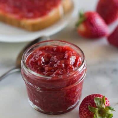 A small mason jar full of strawberry jam made in the instant pot, with fresh strawberries around it and a plate with toast and strawberry jam on it in the background