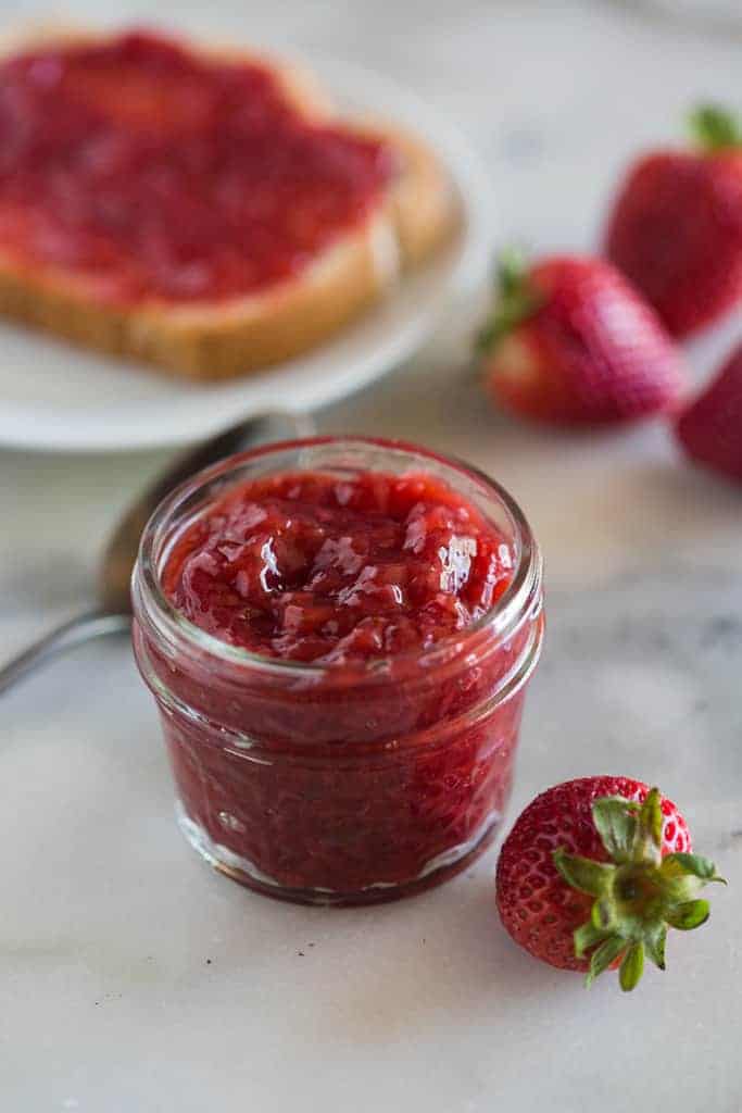 A small mason jar full of strawberry jam made in the instant pot, with fresh strawberries around it and a plate with toast and strawberry jam on it in the background
