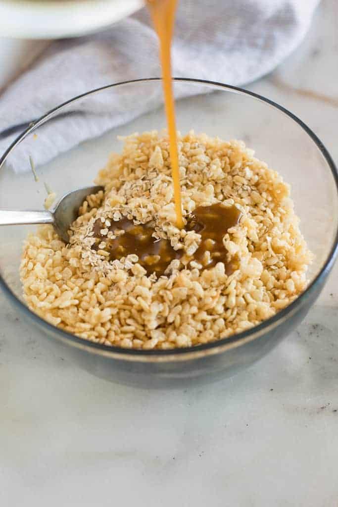 A clear glass mixing bowl with a mixture of quick oats and rice krispies cereal with a syrup being poured over it to make granola bars.