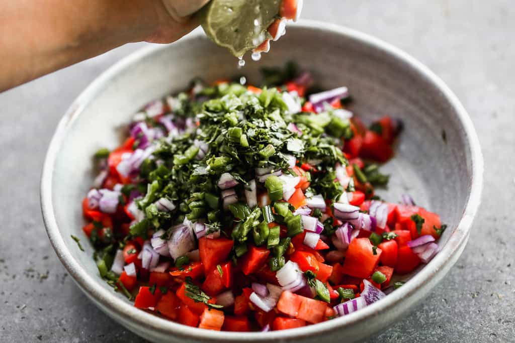 Chopped tomato, onion, cilantro and jalapeño in a bowl with fresh lime juice being squeezed on top.