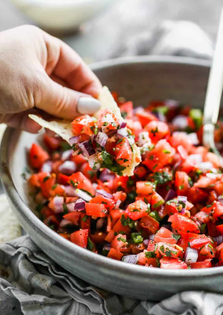 A hand holding a chip, scooping pico de gallo from a bowl.