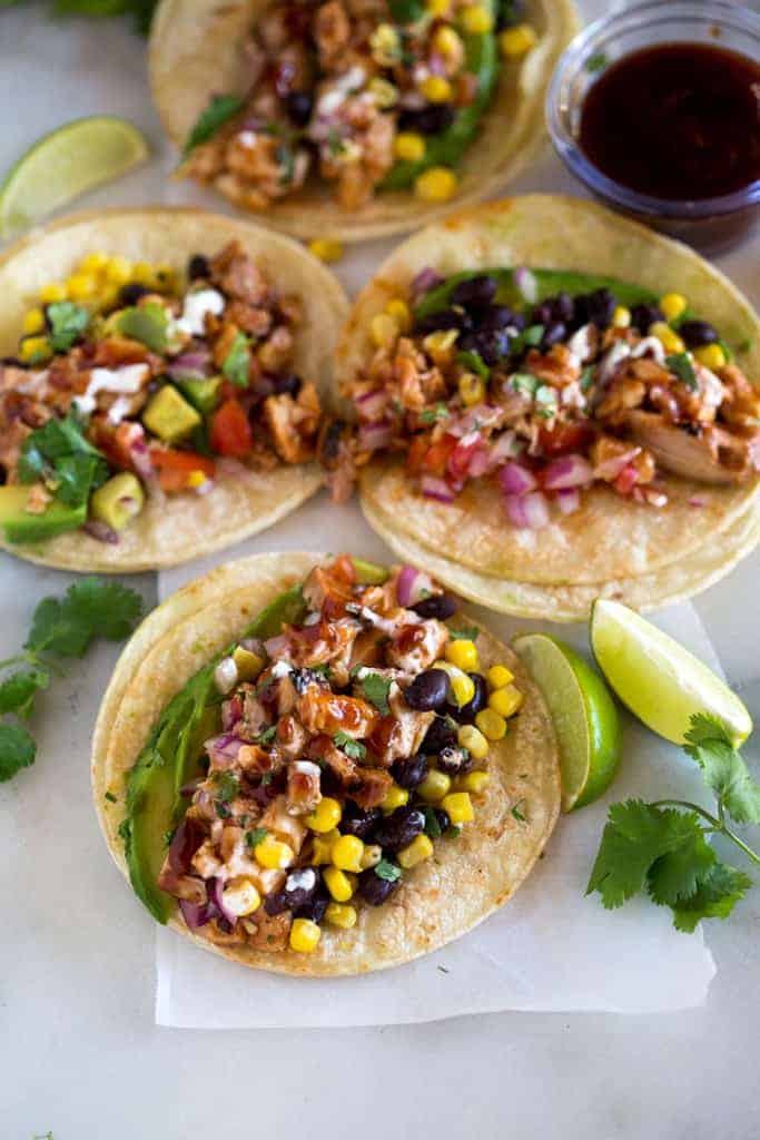 Flat corn tortillas on a white cutting board topped with the ingredients to make bbq chicken tacos including black beans, corn, avocado, cilantro and bbq chicken. 