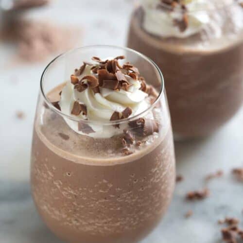 A glass filled with thick frozen hot chocolate, topped with whipped cream and shaved chocolate curls, with a second filled cup in the background.