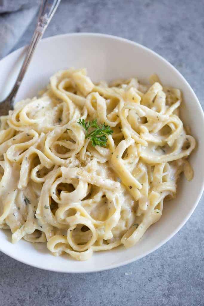 A white pasta bowl full of fettuccine alfredo with a piece of parsley on top for garnish, and a fork on the side.
