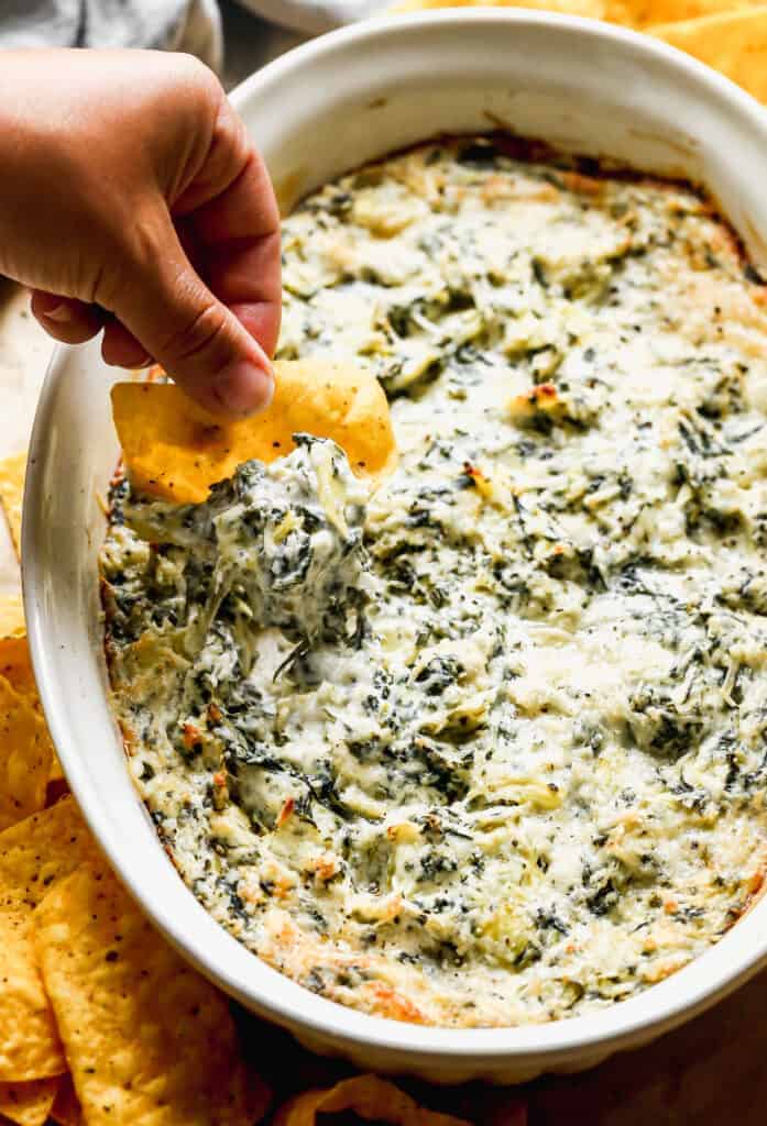 Spinach Artichoke Dip baked in pan, and a hand dipping a chip into it.