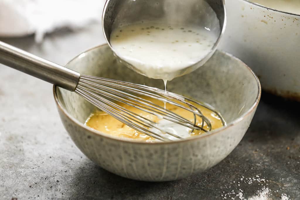 A ladle full of hot tapioca and milk being whisked into a small bowl with beaten eggs, to temper the eggs.