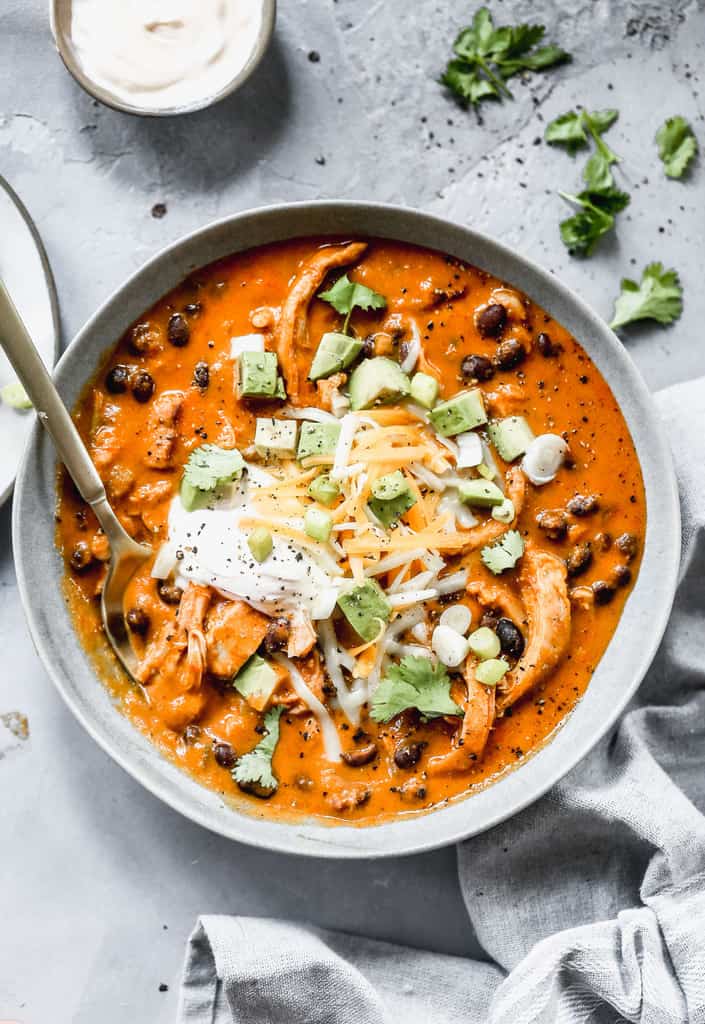 A bowl of chicken enchilada soup, served with sour cream, shredded cheese and chopped avocado on top.