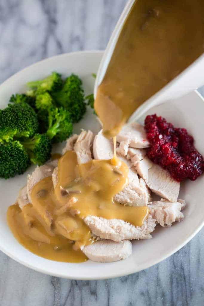 Turkey gravy in a white gravy boat being poured over turkey slices on a plate with cranberry sauce and broccoli.