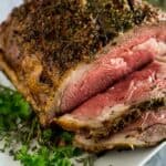 A 5 pound prime rib roast served on a white platter with fresh herbs underneath, with a few slices carved and laying on the platter.
