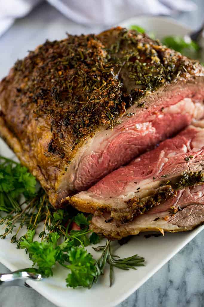 A 5 pound prime rib roast served on a white platter with fresh herbs underneath, with a few slices carved and laying on the platter.