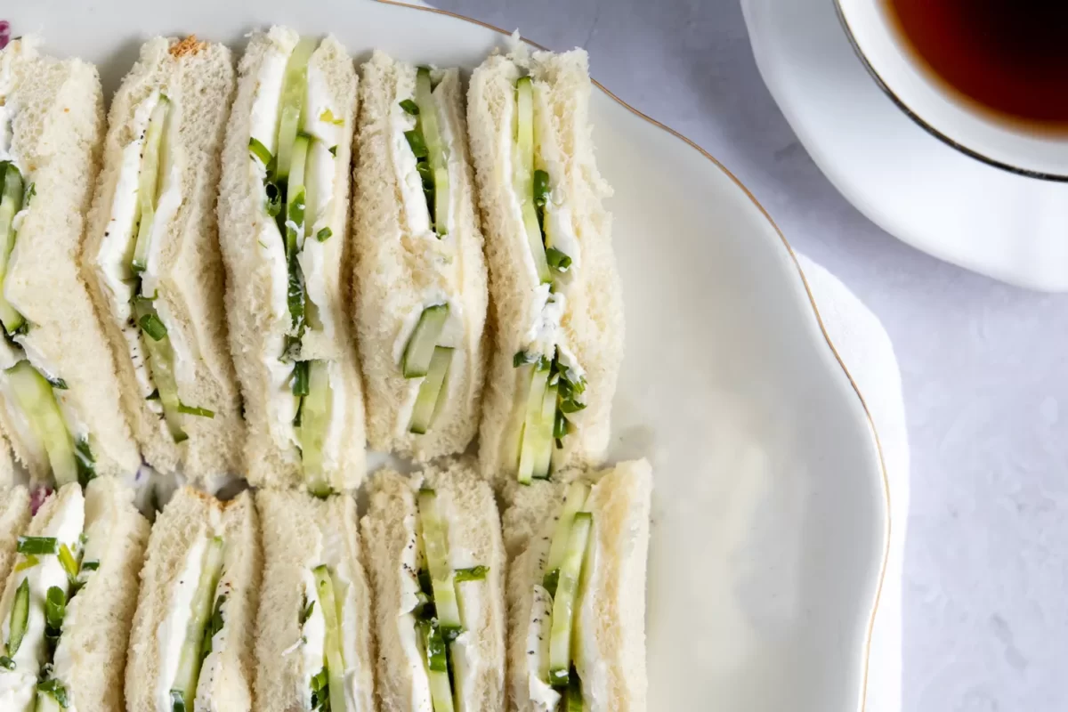 White bread sandwiches filled with cream cheese and cucumber slices lined up on a white platter. 