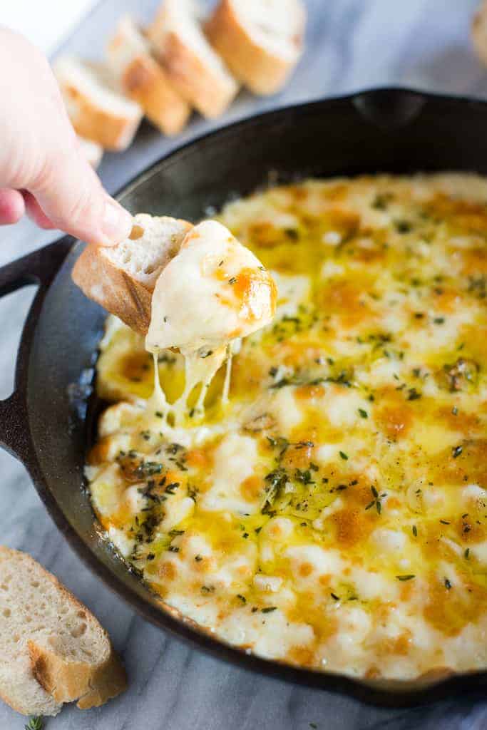 A cast iron skillet with baked fontina cheese dip, bread slices in the background and a hand taking a scoop out of the cheese dip with a bread slice.