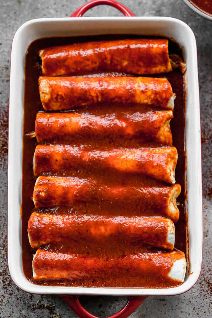 Red enchilada sauce poured over a pan full of filled flour and rolled flour tortillas.