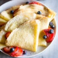 Crepes folded into fourths and sprinkled with powdered sugar, layered on a white plate with sliced strawberries and blueberries.