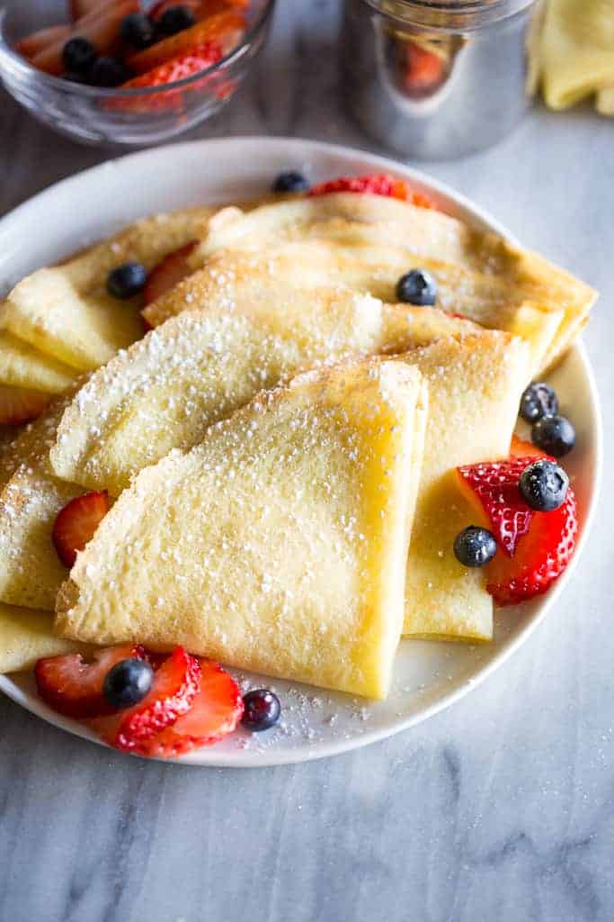 Crepes folded into fourths and sprinkled with powdered sugar, layered on a white plate with sliced strawberries and blueberries.
