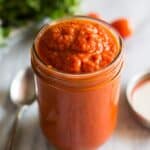 A mason jar filled with red pizza sauce and a spoon and parsley in the background.