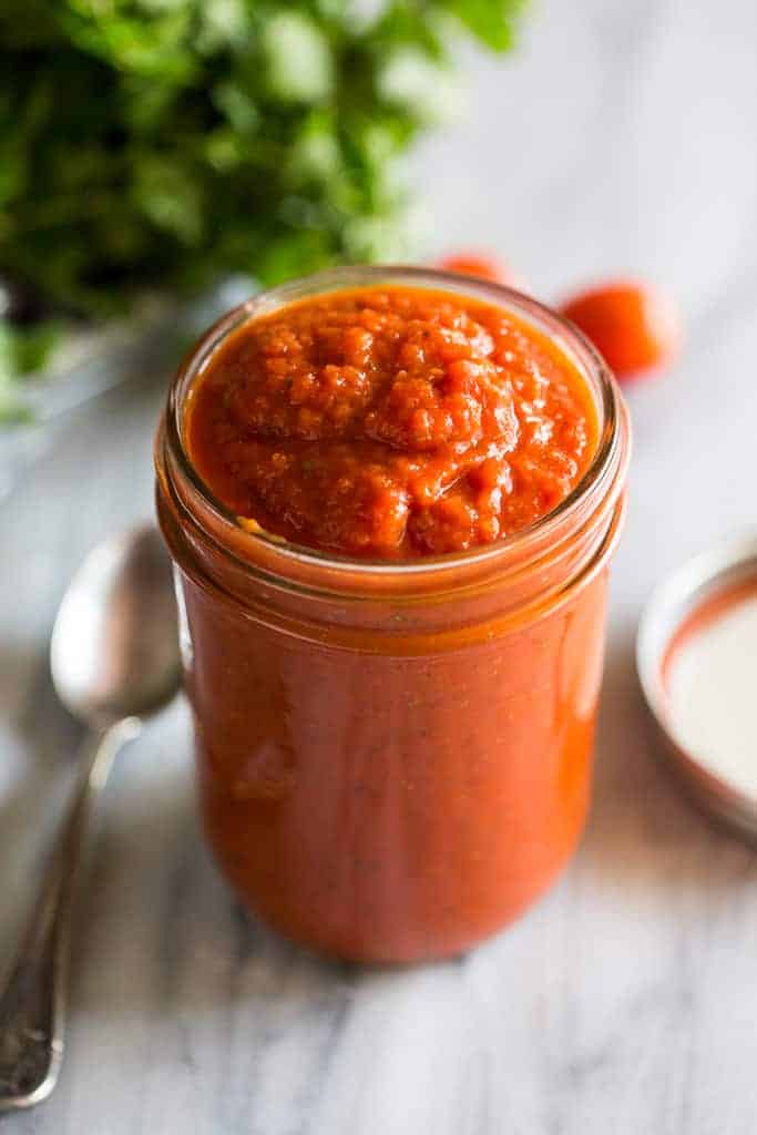 A mason jar filled with red pizza sauce and a spoon and parsley in the background.