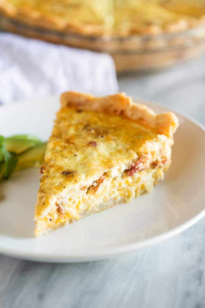 A slice of quiche lorraine on a white plate with sliced avocados on the side.