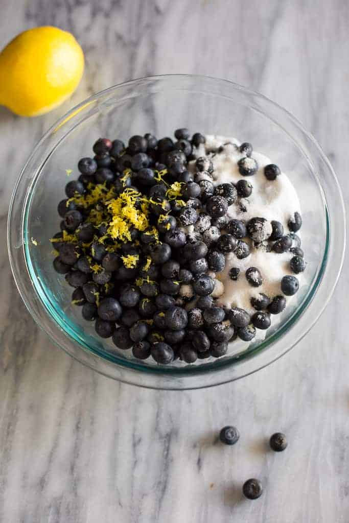 A clear glass bowl with fresh blueberries, sugar and lemon zest to make the filling for blueberry cobbler.