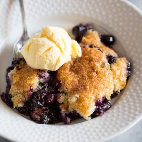 Blueberry cobbler in a white bowl with a spoon and ice cream on top.