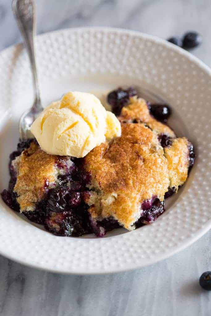 Blueberry cobbler in a white bowl with a spoon and vanilla ice cream on top. 