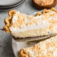 An easy Coconut Cream Pie recipe in a pie dish with one slice being lifted to show the creamy custard layer and fluffy whipped cream and toasted coconut sprinkled on top.