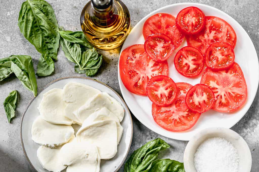 The ingredients needed to make Caprese salad, including fresh mozzarella, basil, tomato and oil.