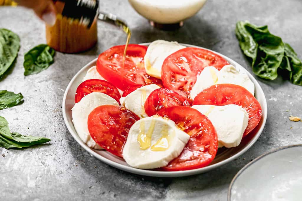 Olive oil being drizzled over a plate of fresh sliced tomato and mozzarella.