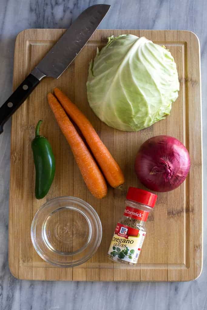 Wood cutting board with ingredients for curtido, including cabbage, carrots, red onion, oregano, vinegar and knife.