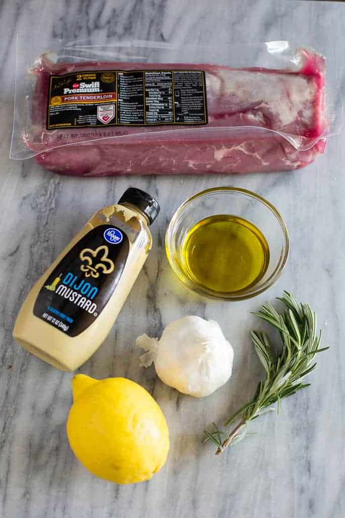 Overhead photo of the ingredients for pork tenderloin marinade including a lemon, garlic cloves, rosemary, olive oil, dijon mustard and a package of pork tenderloins.