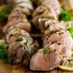 Sliced pork tenderloin on a cutting board, garnished with chopped parsley and lemon slices.