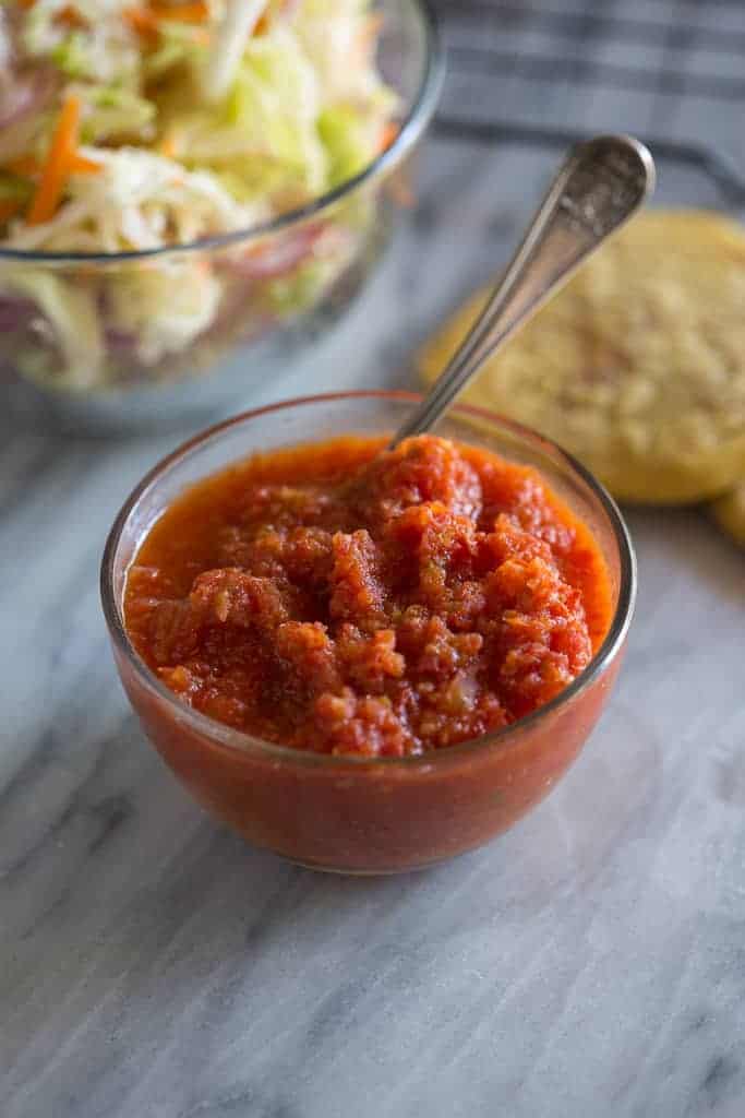 Salsa roja in a glass bowl with a spoon and pupusas and curtido in the background.