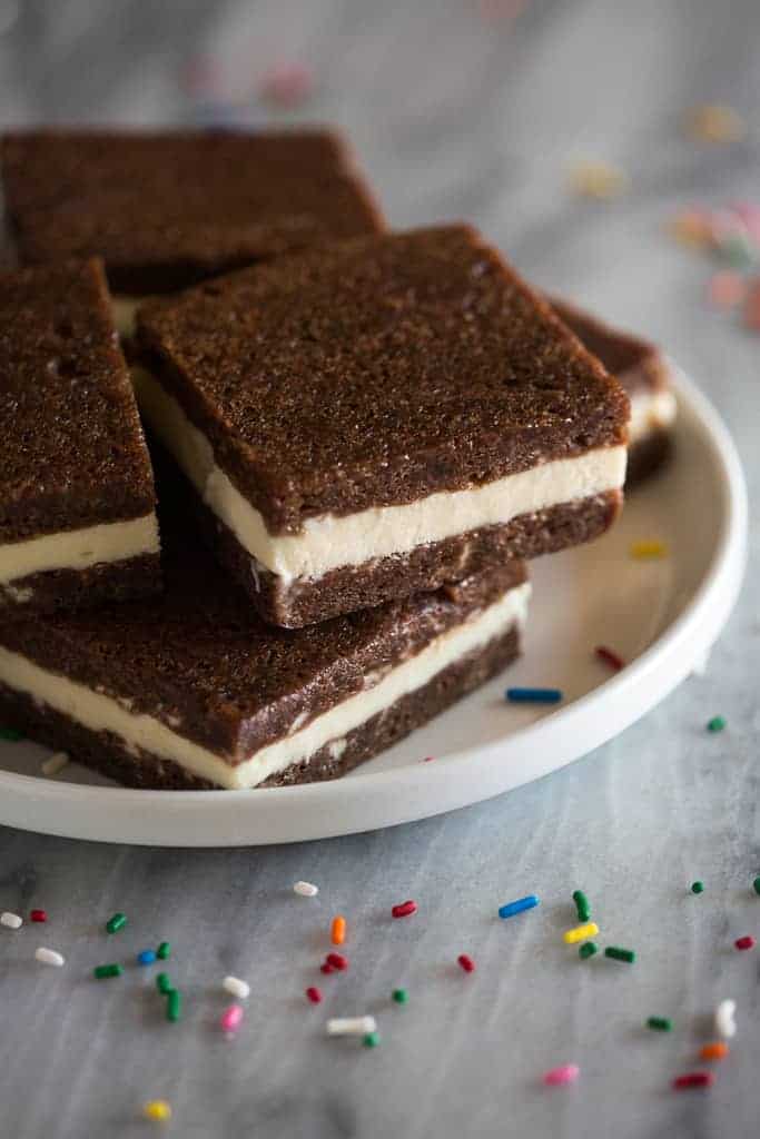 Homemade brownie ice cream sandwich squares stacked on a white plate with sprinkles on the side.