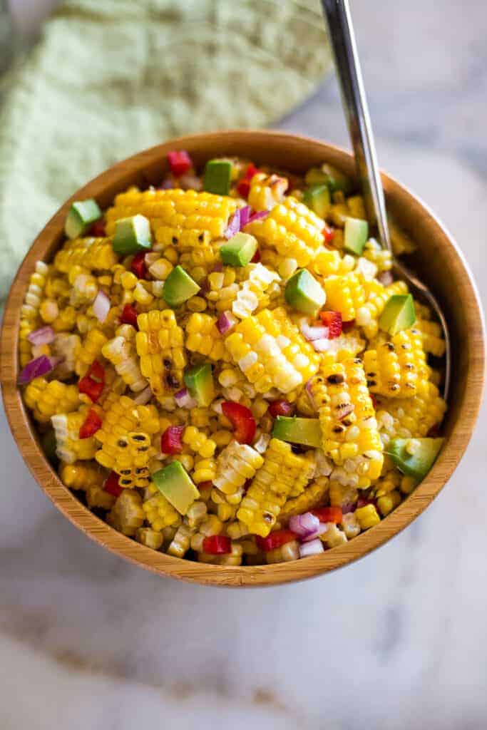 Corn salad made with grilled sweet corn, diced avocado, bell pepper and onion served in a wooden bowl with a spoon.