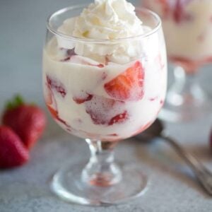 A glass with sliced strawberries and cream sauce in it, another glass and strawberries in the background.