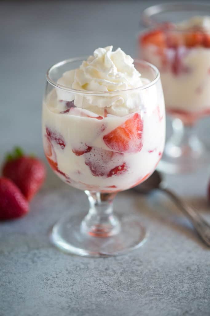 A glass with sliced strawberries and cream sauce in it, another glass and strawberries in the background.