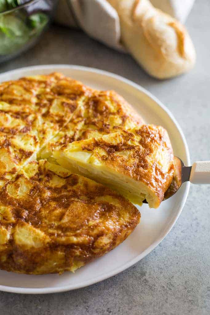 Spanish tortilla de patata on a plate with a slice being served and a baguette and salad in the background.