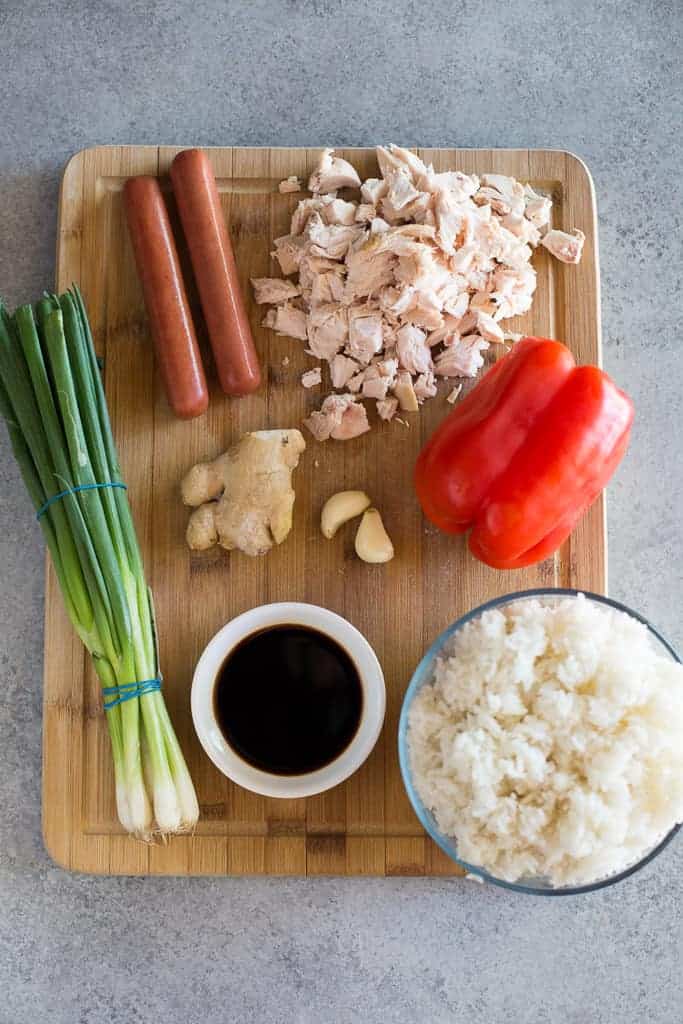 A wood cutting board with the ingredients for arroz chaufa, including green onion, hot dogs, chopped cooked chicken, red bell pepper, ginger, garlic, soy sauce and a bowl of cooked rice.