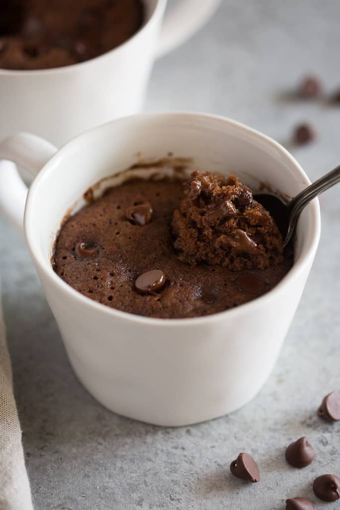 Chocolate cake with chocolate chips microwaved in a white cup and a spoon spooning out a piece.
