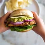 Hands holding the middle of the chicken burger up towards the camera lens, with potato chips on a plate in the background.
