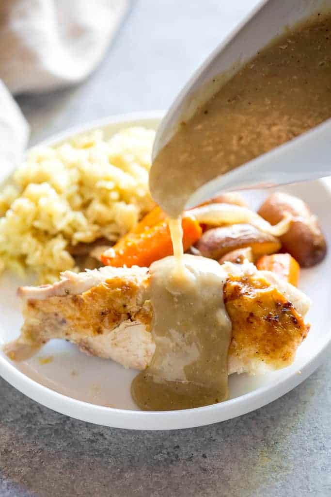 Chicken gravy in a white gravy boat being poured over a chicken breast on a plate with sides of rice and vegetables.