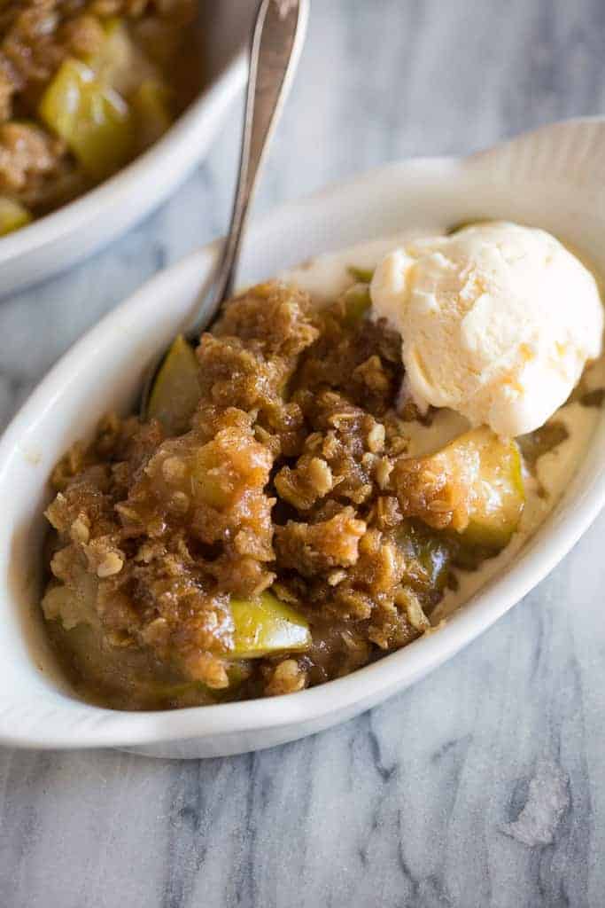 Close up photo of a white dish with apple crisp in it and a scoop of vanilla ice cream on top. 