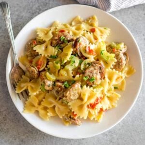 A white bowl with cooked farfalle pasta with sausages and diced red bell pepper with a fork in the bowl.