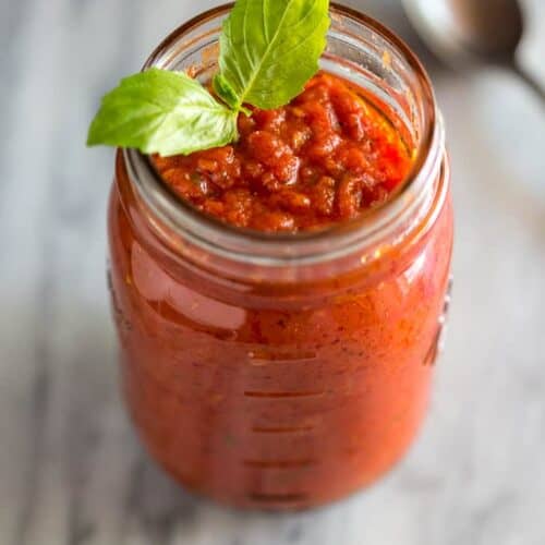 A mason jar full of homemade marinara with a basil leaf on top and spoon to the side.