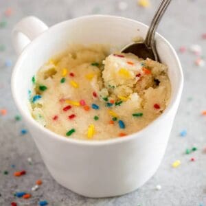 Vanilla cake with sprinkles cooked inside a white mug, with a spoon in it and sprinkles on the board around it.