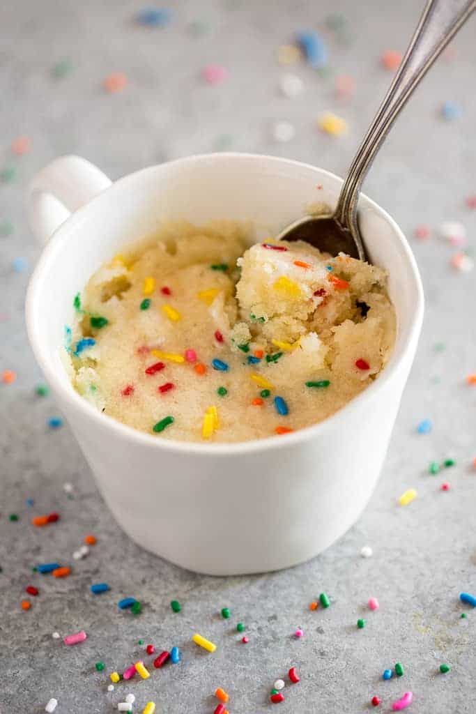 Vanilla cake with sprinkles cooked inside a white mug, with a spoon in it and sprinkles on the board around it.