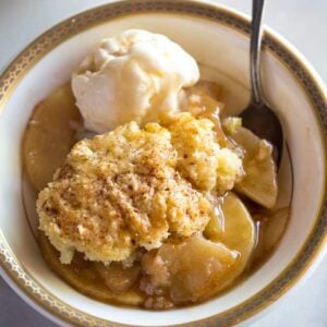 A cream bowl with gold rim full of apple cobbler and a scoop of ice cream.