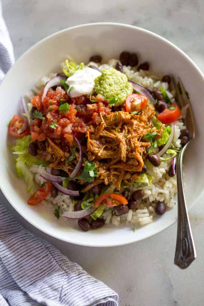 A white bowl with rice, black beans, sweet pork, salsa, guacamole and sour cream.