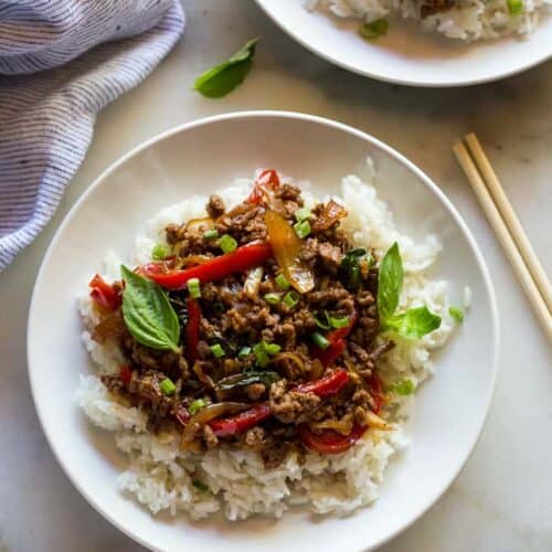 A white plate with white rice and Thai basil beef on it, chopsticks on the side and another plate of food behind it.